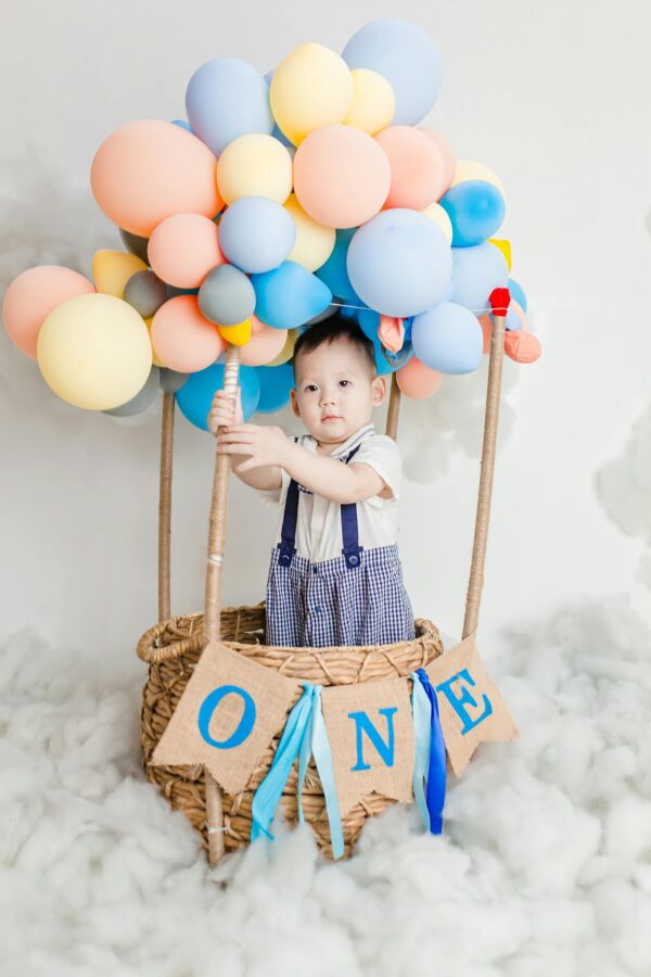 A cute child in a hot air balloon themed setup with colorful balloons and a basket.